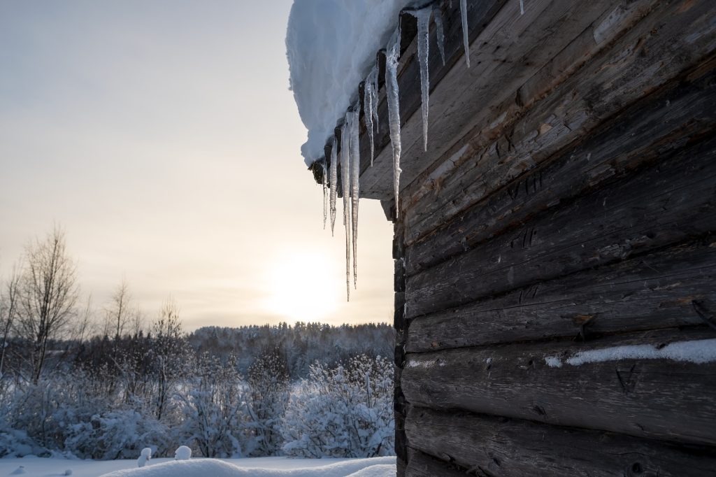 Icicles hang from the snow-covered roof of a log bath, against the landscape, in countryside